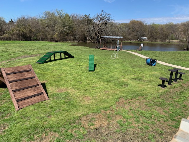 The full dog agility course — ramps, hoop, tunnel, and stepping pads — beside the pond · Waggin' Tail Ranch RV Resort, Pilot Point, Texas